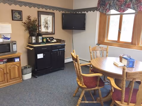 Small dining area with a round wooden table and chairs, a wall-mounted TV, and a coffee/microwave station beneath framed artwork.