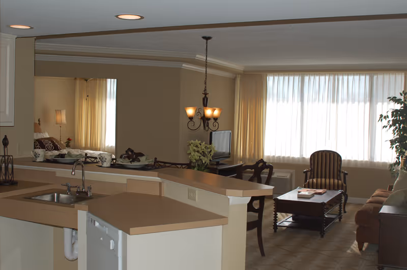 Open-plan kitchen and living area with a sink and breakfast bar opening to a seating area with armchairs, sofa, coffee table and a large curtained window.