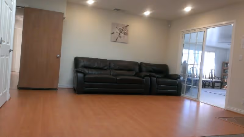 Spacious living room with a black leather sofa against a beige wall, wood flooring, and sliding glass doors opening to a dining area.