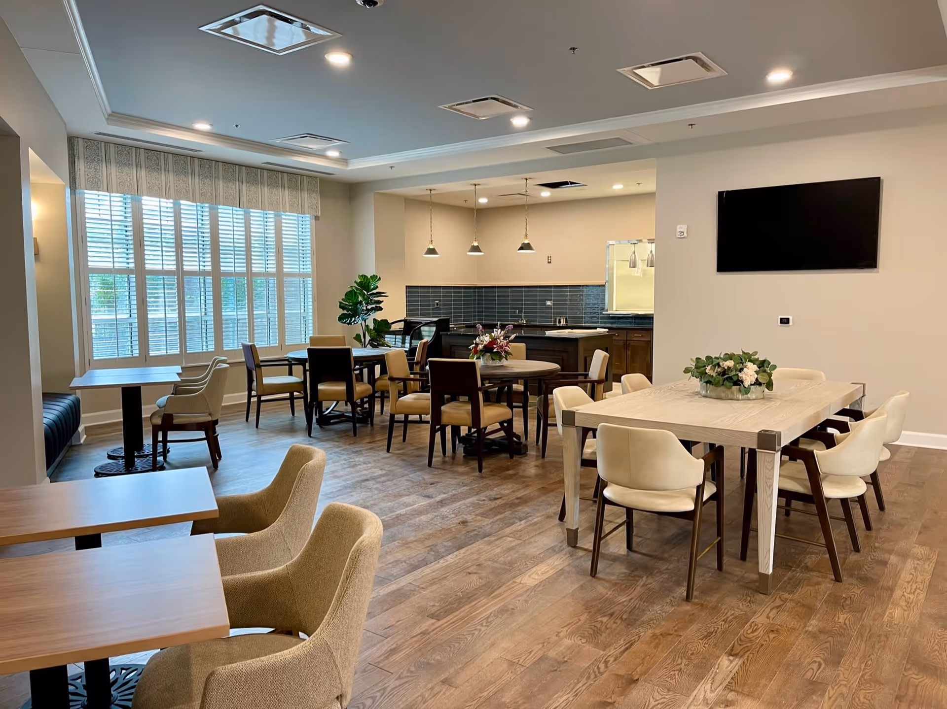 A bright dining area with multiple tables and chairs arranged on a wooden floor. There is a large window with blinds on the left side, a flat-screen TV mounted on the wall to the right, and a small kitchen area with pendant lights in the background. The tables have floral centerpieces, and the room has a modern, clean design.
