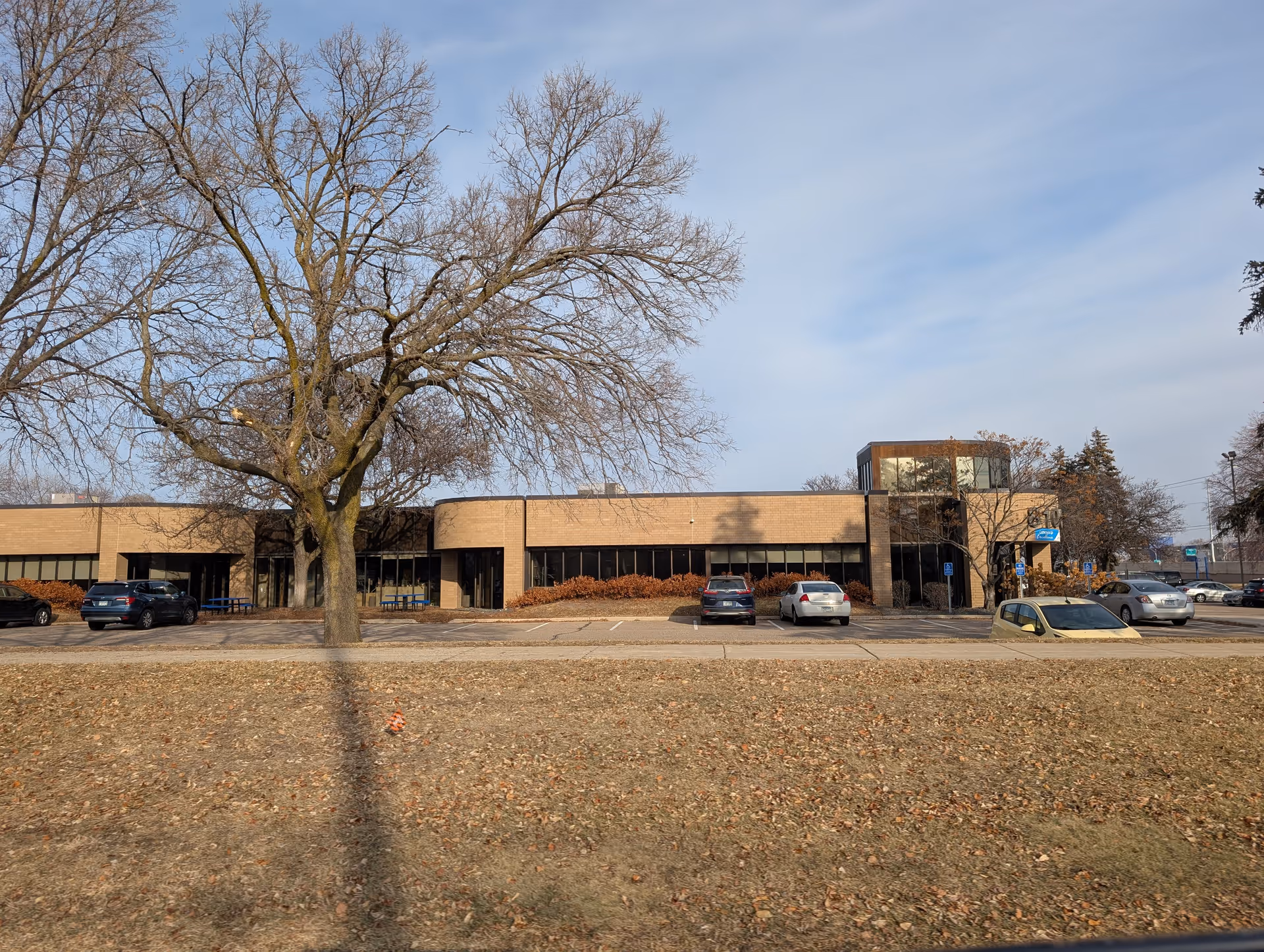 Exterior view of a single-story brick building with large windows and a small parking lot in front. Several cars are parked, and leafless trees are visible around the building under a partly cloudy sky.