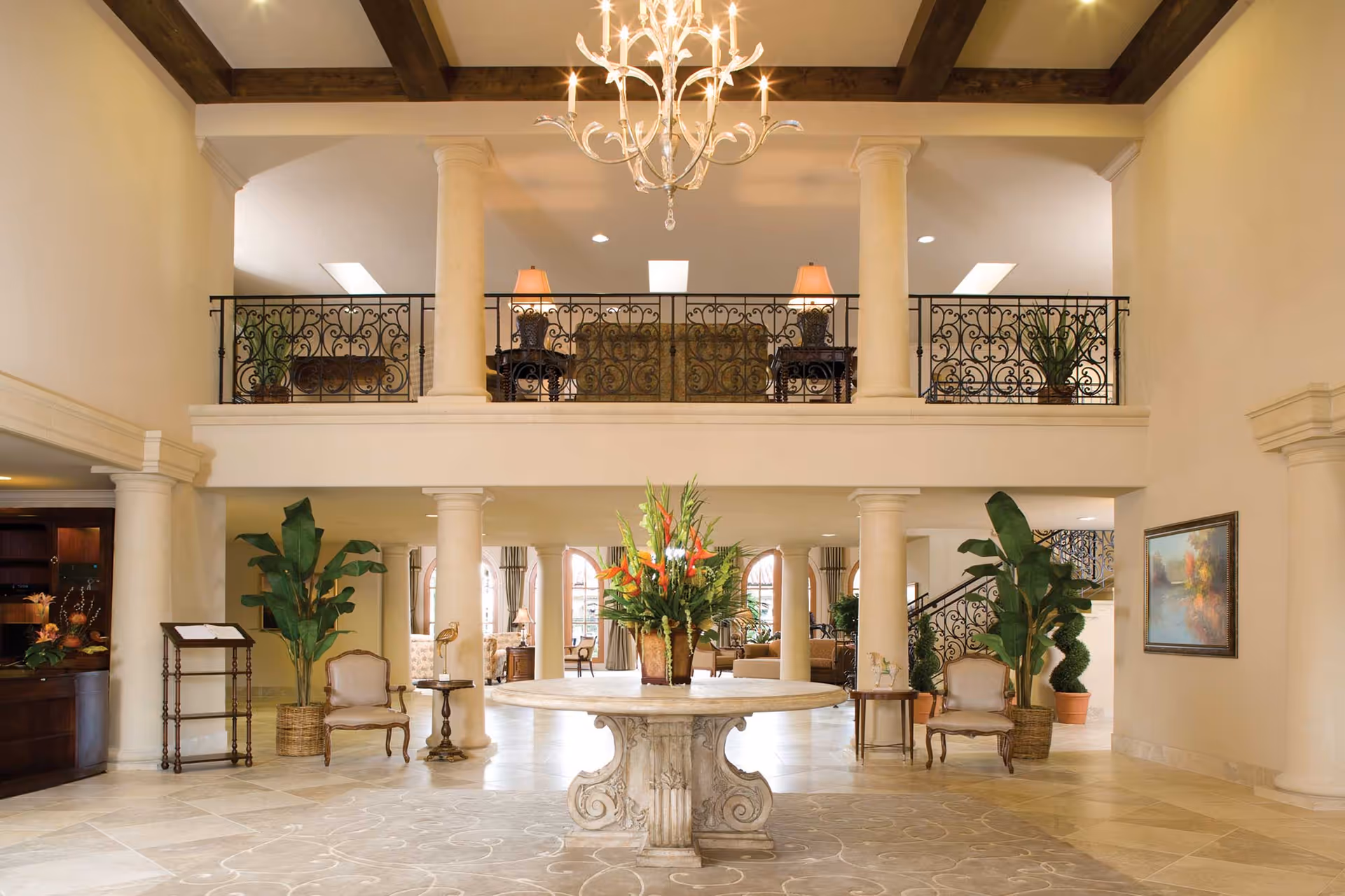 Spacious and elegant interior lobby area with a large round stone table in the center holding a floral arrangement. The room features tall columns, a chandelier hanging from a ceiling with wooden beams, wrought iron railings on the upper balcony, and comfortable seating areas with plants and artwork on the walls.