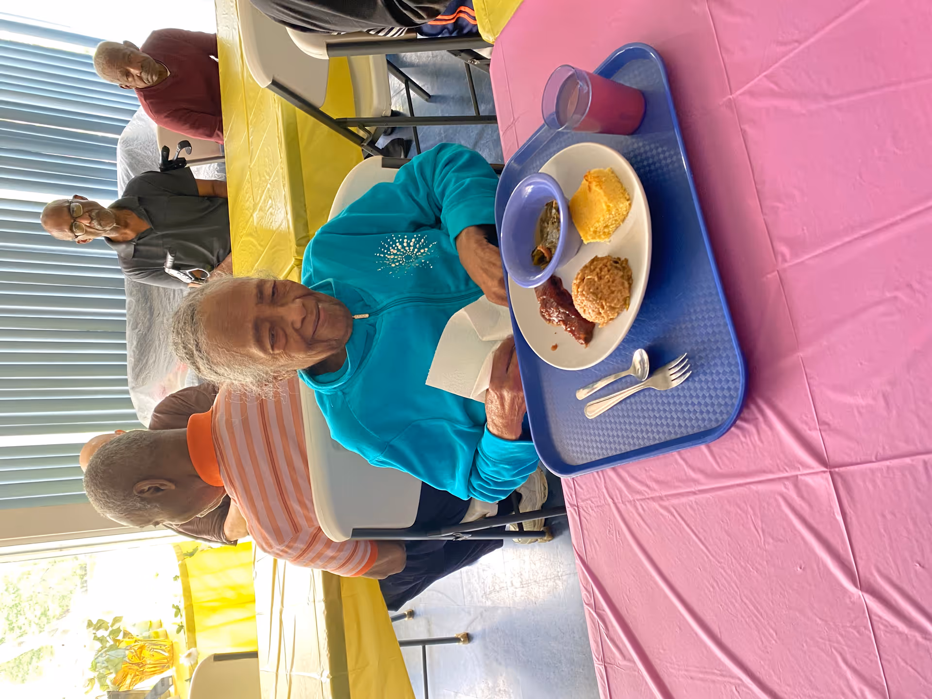 Elderly residents seated at a communal dining table, one woman in a turquoise jacket smiling with a tray of food.