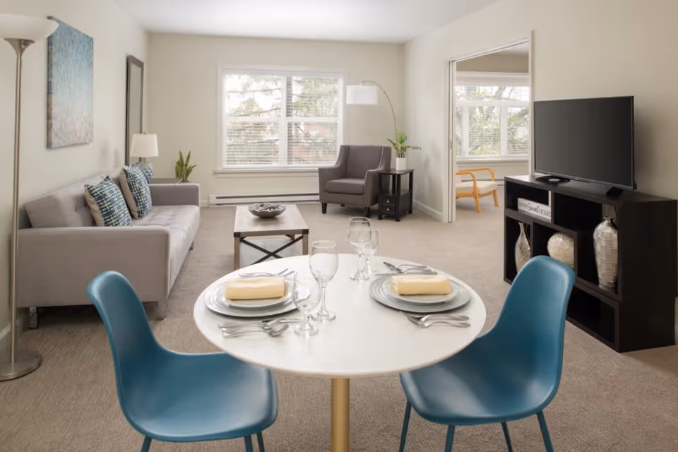A bright living and dining area in a senior living facility with a round white dining table set for two with plates, napkins, and glassware. The room features a gray sofa with patterned cushions, a gray armchair, a coffee table, a floor lamp, and a TV on a dark wooden stand with decorative vases. Large windows let in natural light, and a doorway leads to another room with a wooden chair and more windows.