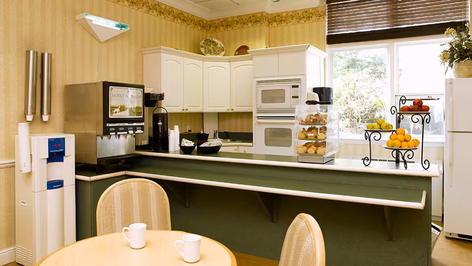 Communal dining/kitchen area with a service counter, beverage machines, pastry display, fruit stand, and a small table with two mugs.