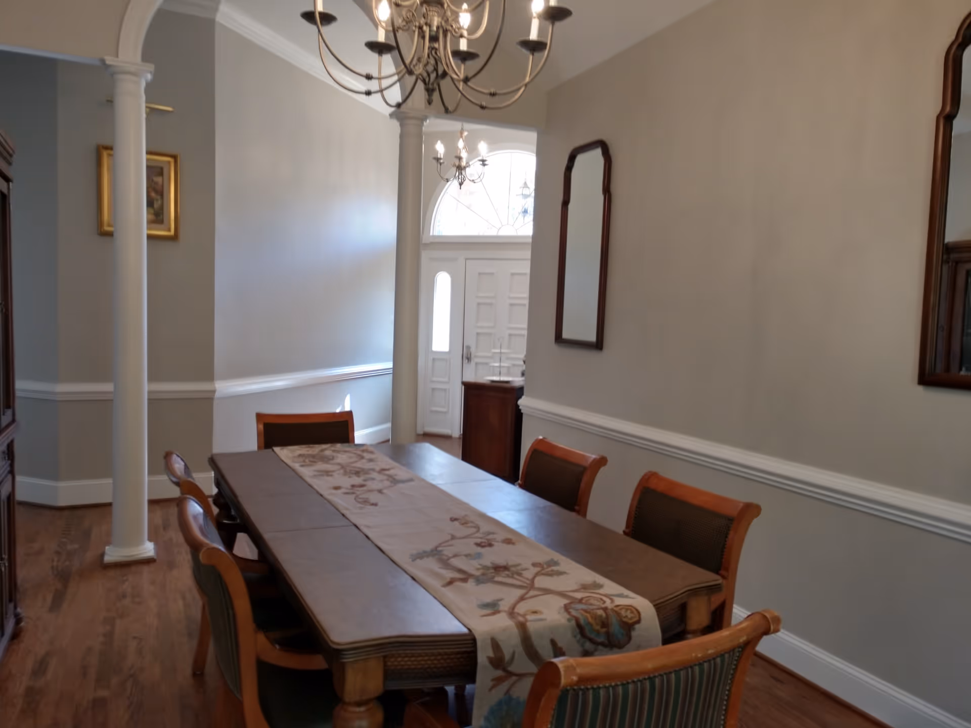 A dining room with a wooden dining table covered by a decorative table runner and surrounded by six chairs. The room features hardwood flooring, two white columns, a chandelier hanging from the ceiling, and a large windowed front door in the background. There are two mirrors on the right wall and a framed picture on the left wall.