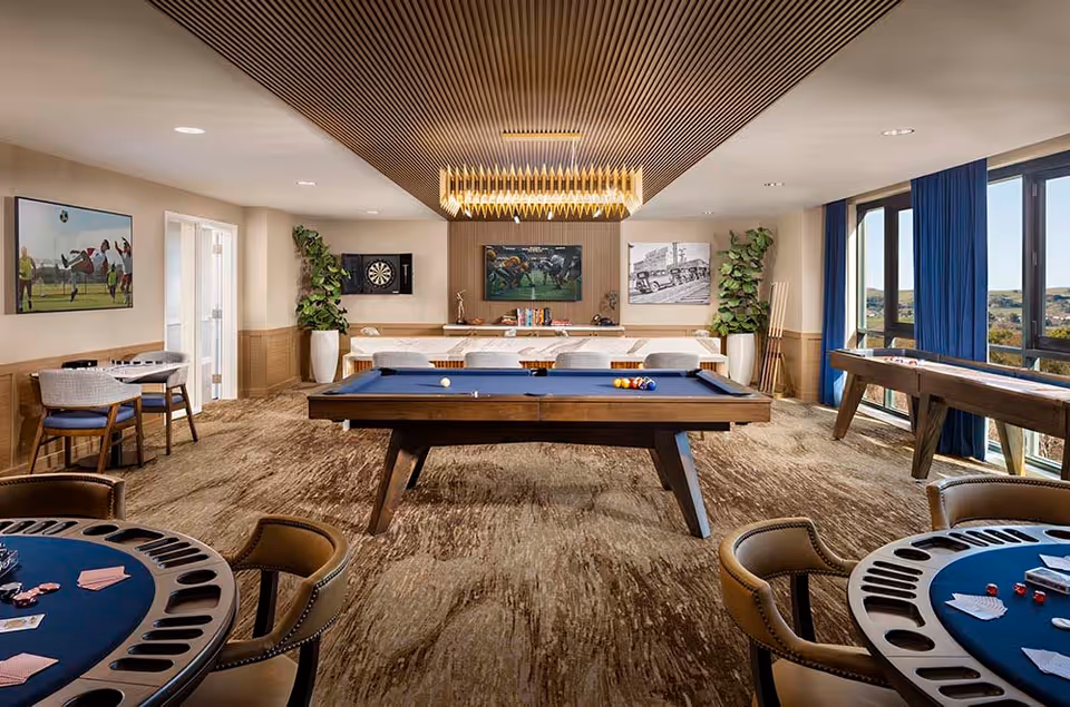 A spacious game room with a pool table in the center, surrounded by chairs and tables for card games. The room features large windows with blue curtains, a dartboard on the wall, and various framed pictures. There are also plants and a shuffleboard table near the windows.