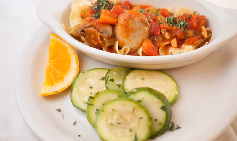 A white plate with a serving of pasta topped with tomato sauce and mushrooms, garnished with herbs. On the side, there are several slices of cucumber sprinkled with herbs and a wedge of orange.