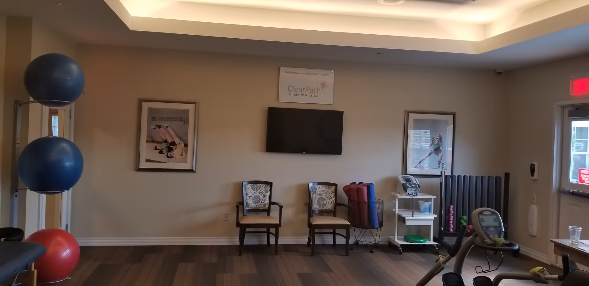 A small exercise room with two large blue exercise balls and one red exercise ball on a rack to the left. Two chairs with floral cushions are placed against the wall beneath a mounted flat-screen TV and a ClearPath Home Health & Hospice sign. There are framed pictures on the wall, exercise mats rolled up in a basket, and various exercise equipment including a stationary bike and weights. A door with a red exit sign is visible on the right side.