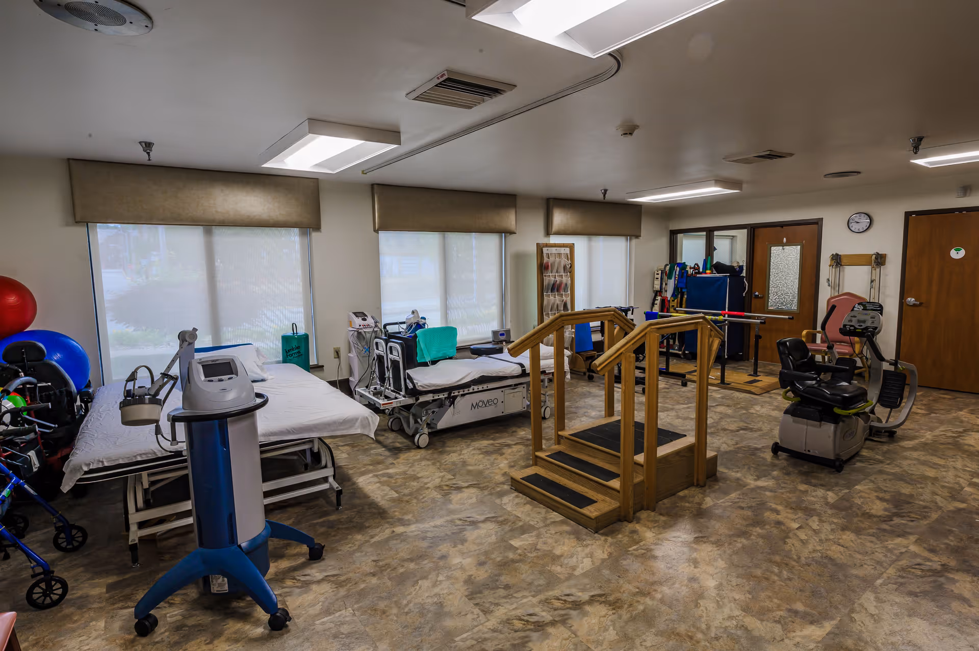 A rehabilitation therapy room with medical beds, exercise equipment including a stationary bike, parallel bars with wooden handrails for walking practice, therapy balls, and other physical therapy tools. The room has large windows with blinds, beige walls, and a tiled floor.