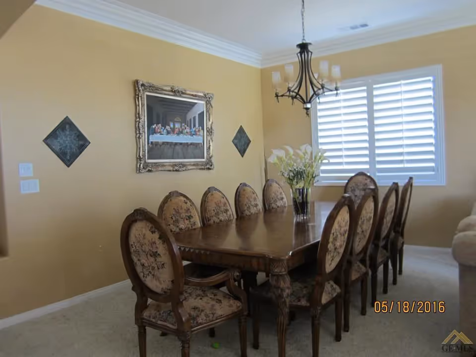 A dining room with a long wooden table surrounded by eight upholstered chairs with floral patterns. A vase with white flowers is placed in the center of the table. The walls are painted yellow and decorated with a framed painting and two diamond-shaped wall hangings. A chandelier with multiple lights hangs above the table, and a window with white shutters is visible in the background.