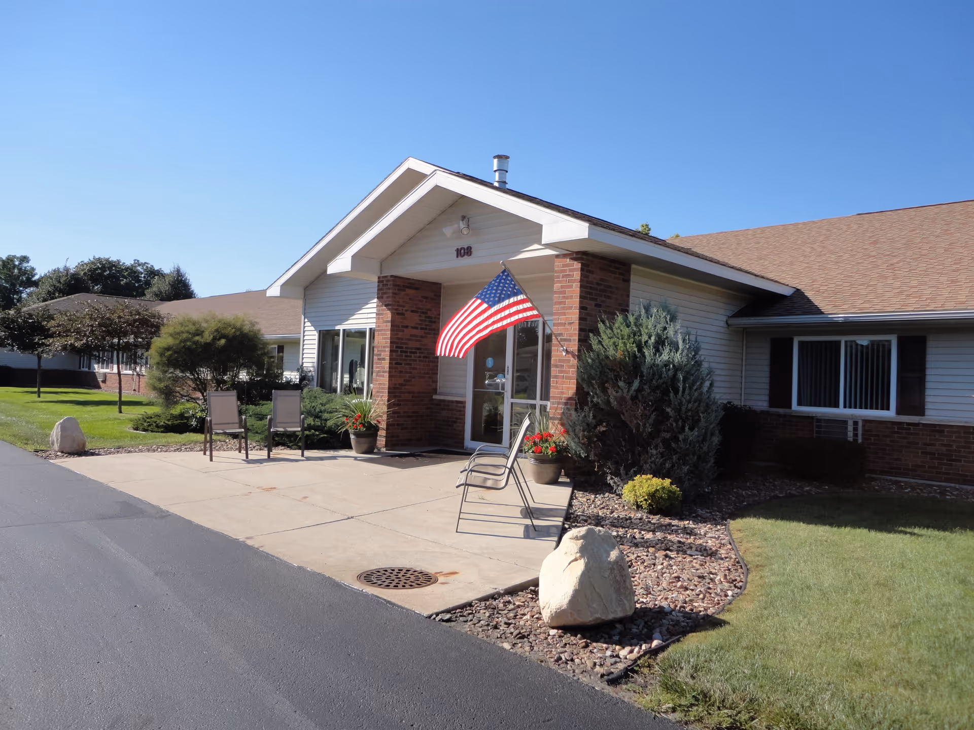 Front entrance of a single-story senior living building with a covered portico, American flag, potted plants, chairs, and landscaping.