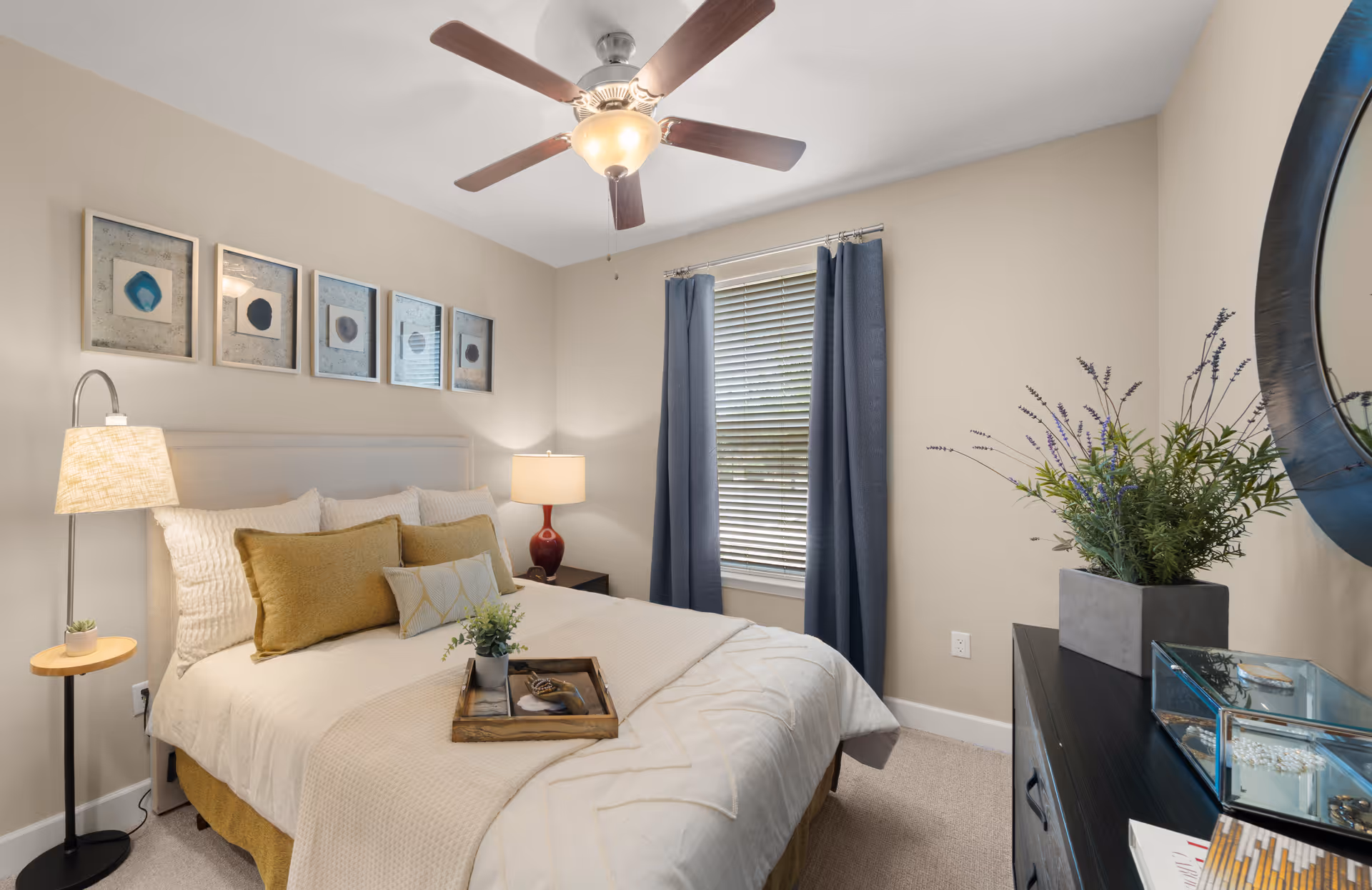 A cozy bedroom with a neatly made bed featuring white and mustard yellow pillows and a beige blanket. There is a wooden tray with a plant and a book on the bed. The room has a ceiling fan with a light, a floor lamp, a bedside table with a lamp, blue curtains on the window, and a dresser with a round mirror and a potted plant.