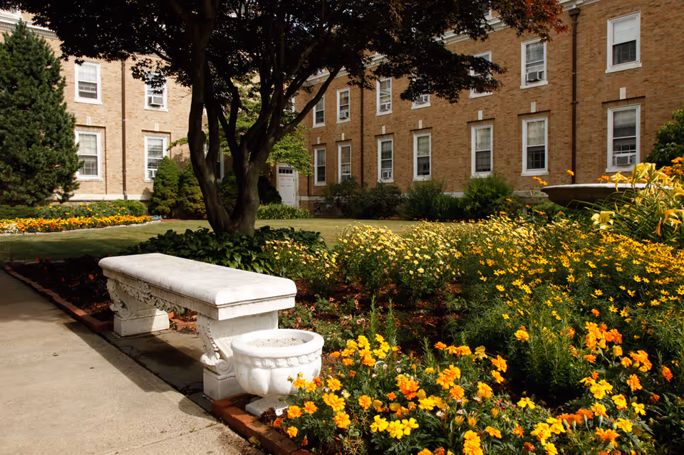 A landscaped courtyard with a white stone bench, flower beds of yellow and orange blooms, and a brick building in the background.