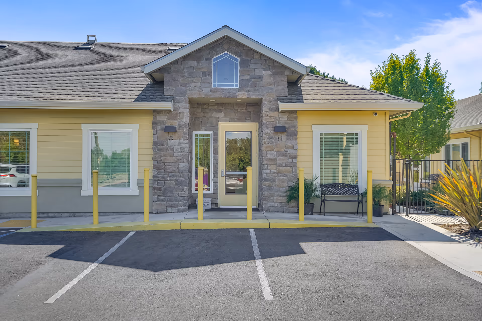 Front exterior view of a single-story building with a stone facade around the entrance door, yellow siding, and a gray shingled roof. There are two windows with white trim on either side of the door, yellow bollards in front of the entrance, a bench to the right, and some greenery including a tree and potted plants. The sky is clear and blue.