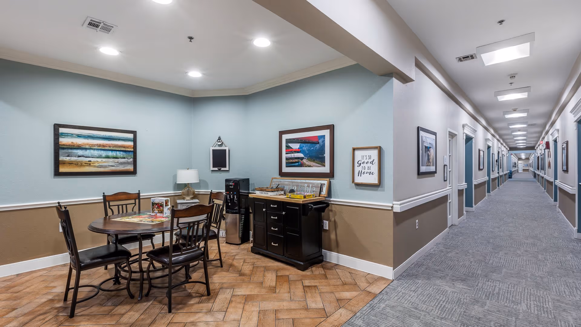 A seating area in a senior living facility with a round table and four chairs on a wood-patterned floor. The walls are painted light blue and beige with white trim. There are framed pictures and a sign that reads 'It's so good to be home' on the walls. A black cabinet with a water dispenser and some items on top is against the wall. To the right is a long, well-lit hallway with multiple doors and framed pictures along the walls.
