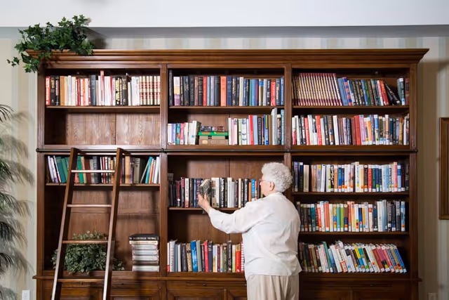 An elderly woman with white hair and glasses is standing in front of a large wooden bookshelf filled with books. She is reaching to place or take a book from the middle shelf. A wooden ladder leans against the bookshelf, and there are some green plants on top and inside the bookshelf.