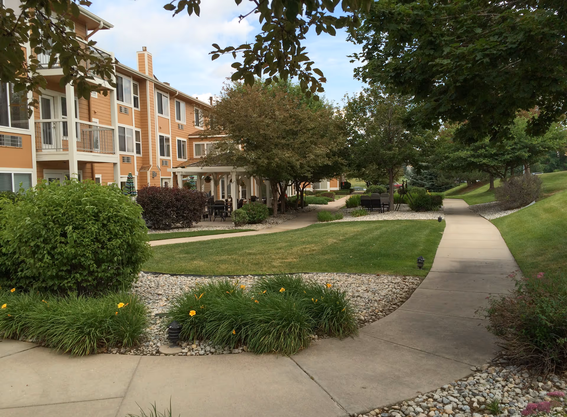 Outdoor view of a senior living facility with a paved walkway winding through a landscaped garden area featuring green grass, bushes, trees, and flower beds. The building has multiple floors with balconies and windows, and there are outdoor seating areas with tables and chairs under a pergola.