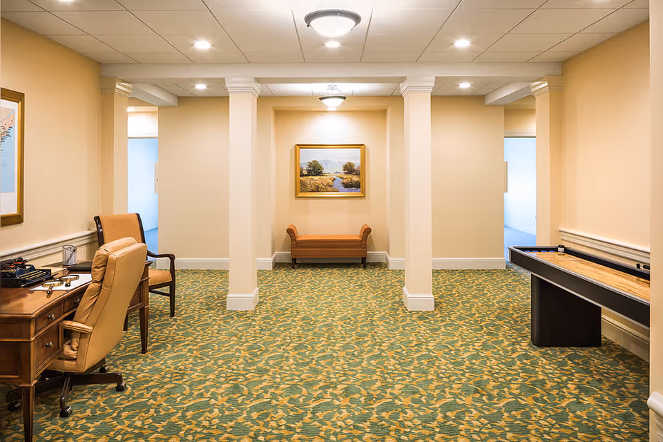 Spacious carpeted common room with columns, a desk and chair, bench, wall painting, and a shuffleboard table.