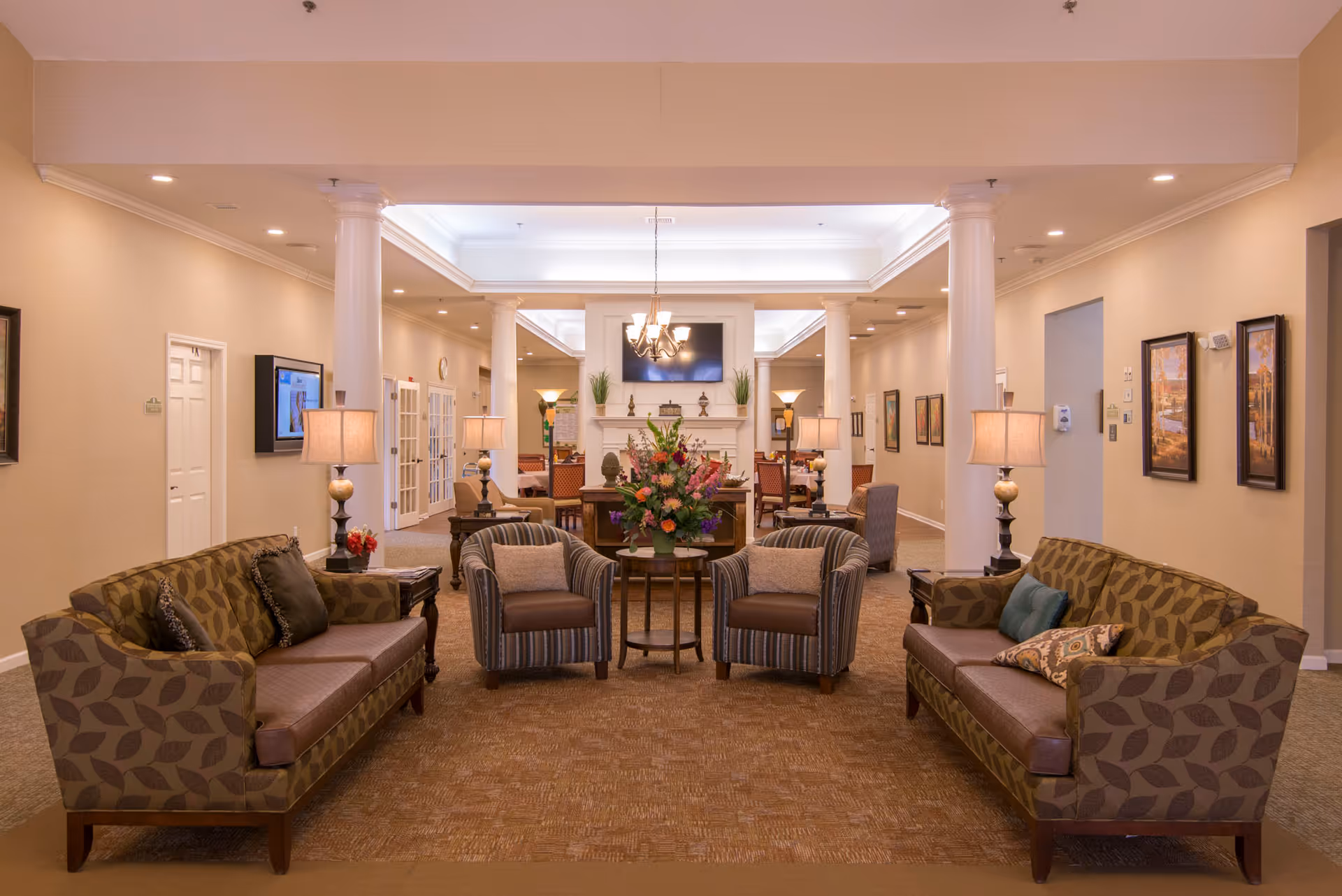 A spacious and well-lit senior living facility common area with two patterned sofas facing each other, two striped armchairs with cushions, a round table with a large floral arrangement in the center, and multiple lamps. The room features beige walls, carpeted floors, white columns, framed artwork on the walls, and a chandelier hanging from the ceiling. In the background, there is a fireplace with decorative items and a mounted flat-screen TV above it.