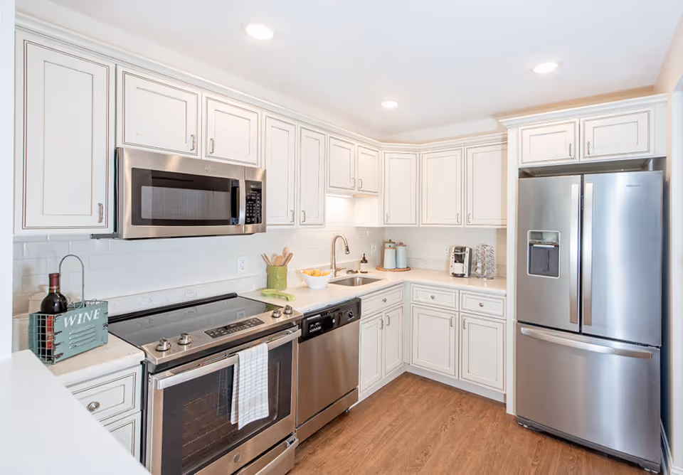 Bright and modern kitchen with white cabinetry, stainless steel appliances including a microwave, stove, dishwasher, and refrigerator. The countertop has a bowl of lemons, a green utensil holder, a coffee maker, and a wine bottle in a decorative holder. The floor is wooden and the space is well-lit with recessed ceiling lights.