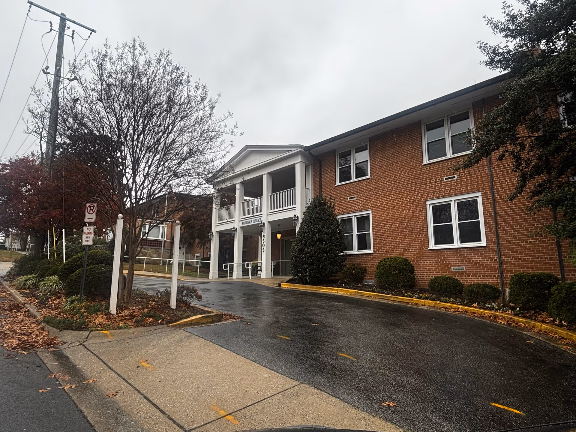 Brick two-story building with a columned portico entrance and a wet driveway under an overcast sky.
