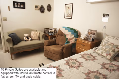 A cozy nursing home room with a bed covered in a patterned quilt, a brown recliner chair with a colorful knitted blanket draped over it, a small green loveseat with pillows and a teddy bear, a wooden nightstand with framed photos and a lamp, and wall decorations including framed pictures and ornamental plates.