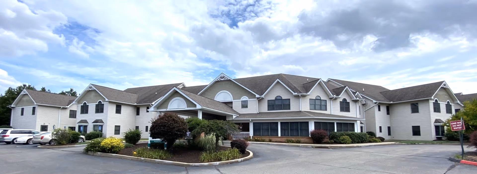 Wide exterior view of a large two-story senior living facility building with beige siding and multiple gabled roofs under a partly cloudy sky. There is a parking lot with several cars on the left side and landscaped bushes and trees in front of the building. A sign near the driveway reads 'ONE WAY DO NOT ENTER.'