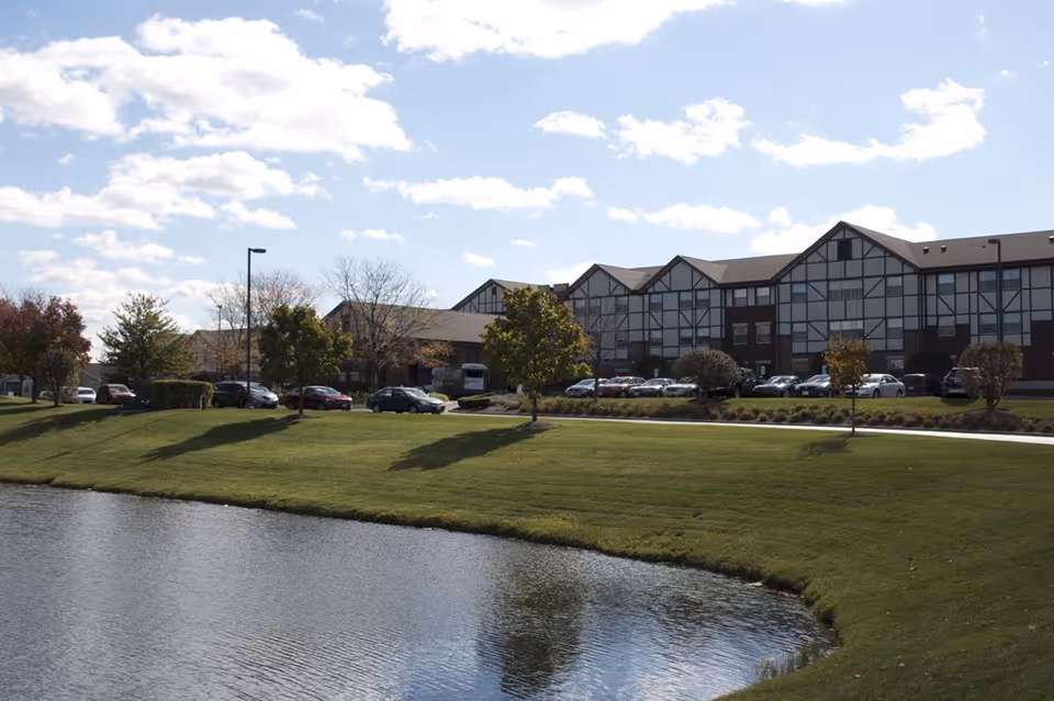 Exterior view of a senior living facility building with Tudor-style architecture, a parking lot with cars, green lawn, trees, and a pond in the foreground under a partly cloudy sky.