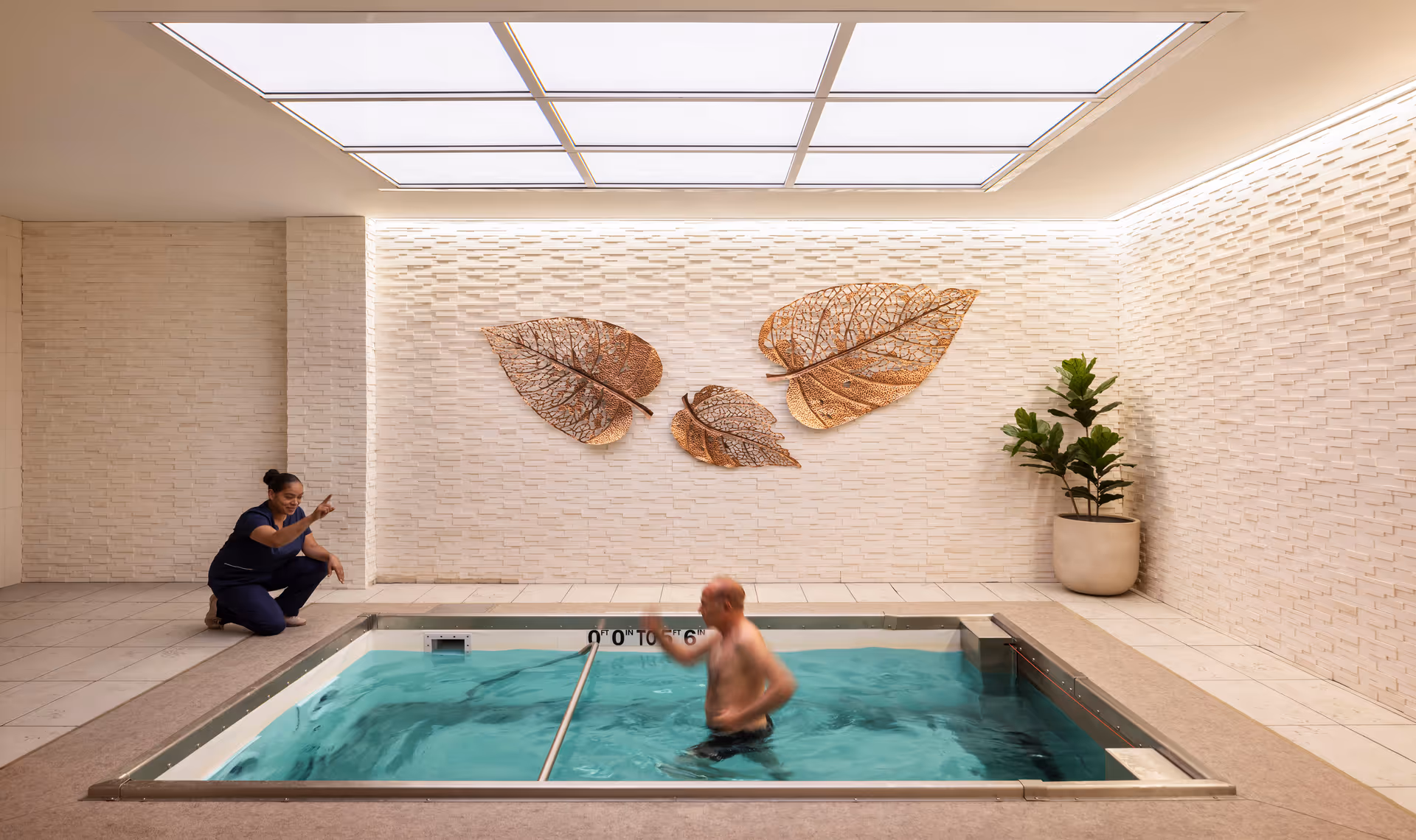 An elderly man is exercising in a small indoor pool while a staff member in navy scrubs kneels beside the pool, encouraging him. The room has beige tiled walls with three large decorative metal leaves mounted on one wall and a potted plant in the corner. The ceiling features large, bright skylights.