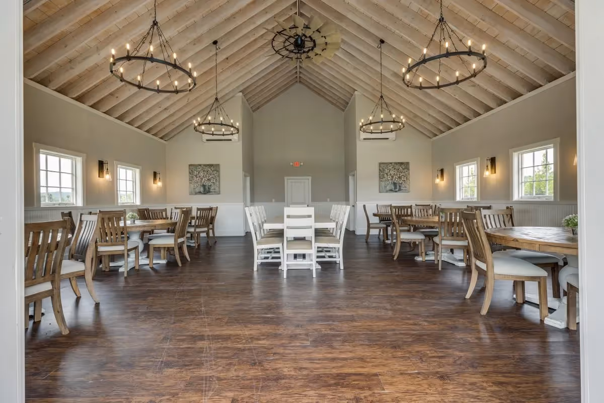 Spacious dining room with wooden floors, multiple round and rectangular wooden tables surrounded by chairs, beige walls with windows, wall-mounted lights, and three large circular chandeliers hanging from a vaulted wooden ceiling.