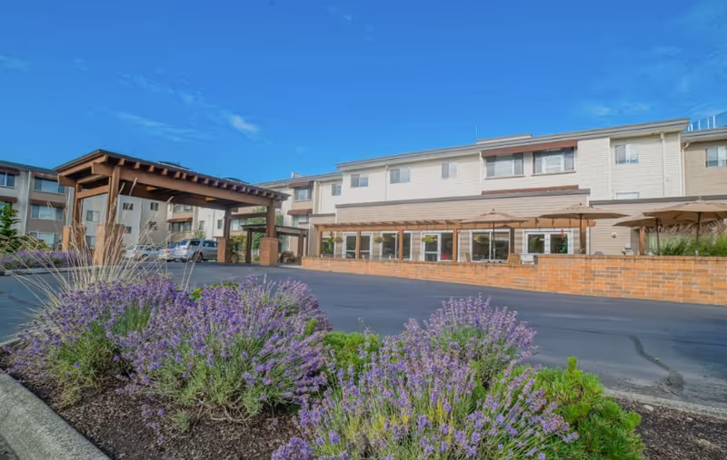 Exterior view of Village Concepts of Auburn - Brannan Park showing a multi-story building with a covered entrance, a paved driveway, and landscaped flower beds with purple flowers in the foreground under a clear blue sky.