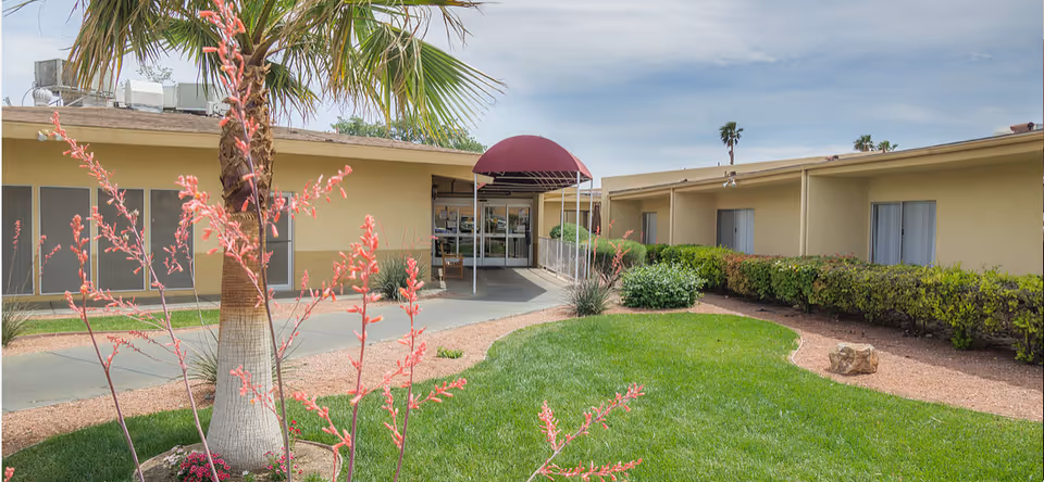 Outdoor view of a senior living facility with a walkway leading to a building entrance covered by a maroon awning. The area features green grass, bushes, a palm tree, and flowering plants under a partly cloudy sky.