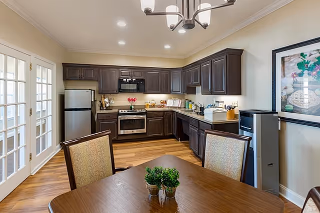 A modern kitchen with dark wood cabinets, stainless steel refrigerator and oven, a wooden dining table with two upholstered chairs, and a small green plant centerpiece. The room has wooden flooring, recessed ceiling lights, and a framed floral picture on the wall.