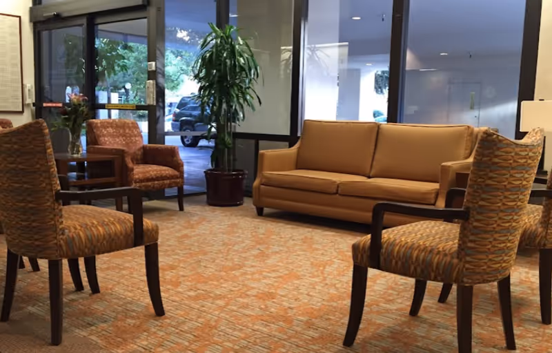 A seating area inside Webster House featuring a tan leather sofa, four patterned armchairs, a potted plant, and a glass door leading outside with a view of parked cars.