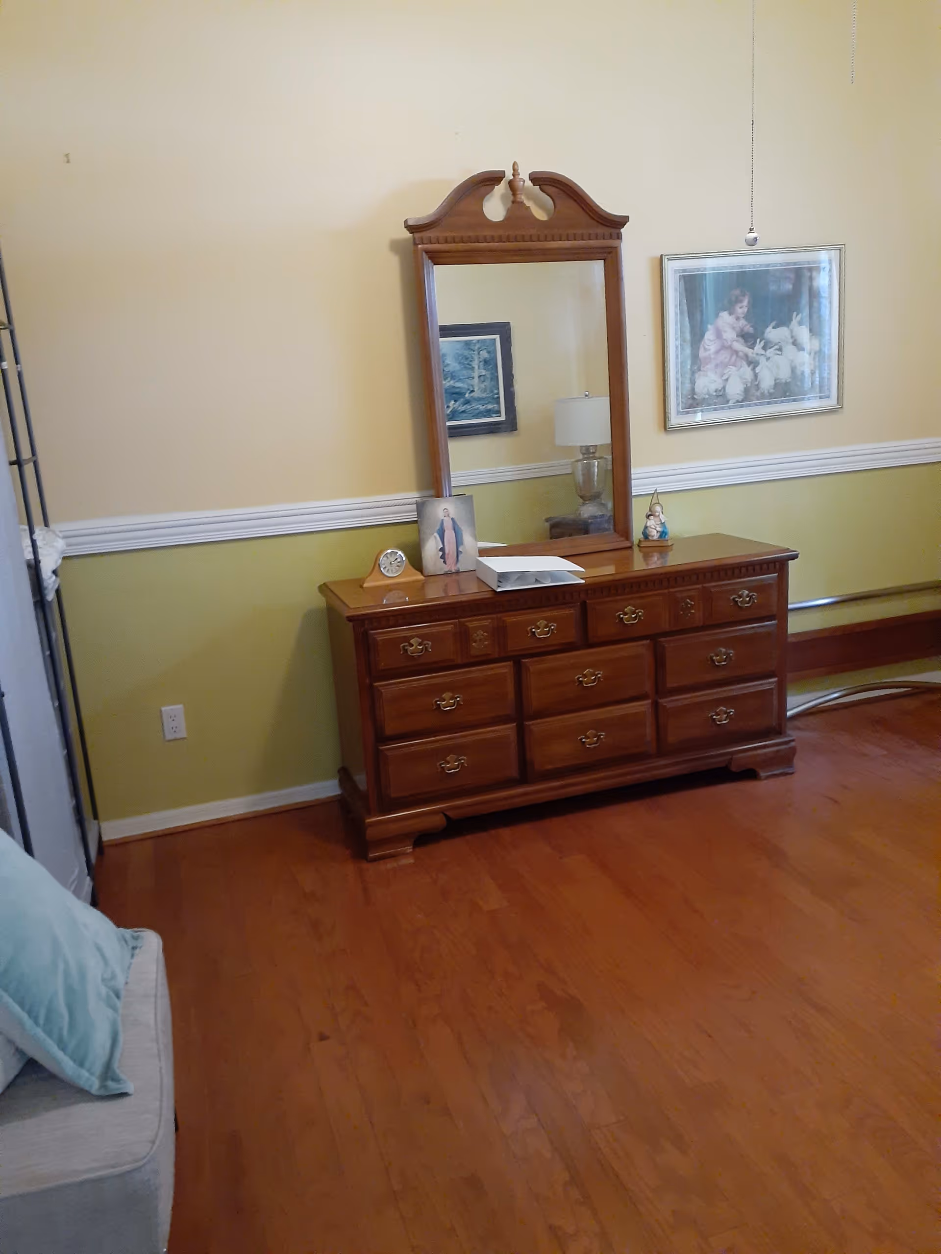 A wooden dresser with multiple drawers and an attached mirror in a room with light yellow and green walls. On top of the dresser are a small clock, a religious picture, a white box, and a small figurine. A framed picture of a child with rabbits hangs on the wall to the right. Part of a light gray couch with a blue pillow is visible on the left side of the image. The floor is wooden.