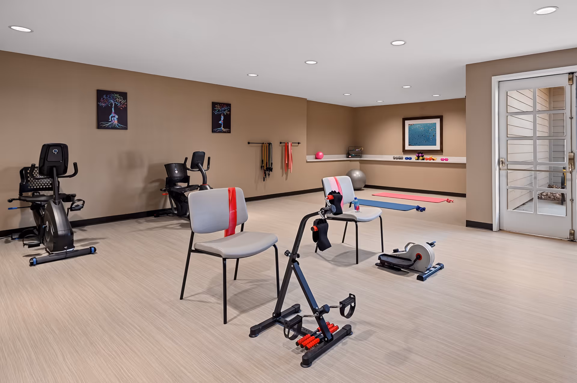 A fitness room with exercise equipment including two stationary bikes, two chairs with resistance bands, a pedal exerciser, yoga mats, dumbbells, and exercise balls. The room has beige walls, light wood flooring, recessed ceiling lights, and a glass door leading outside. There are three framed artworks on the walls.