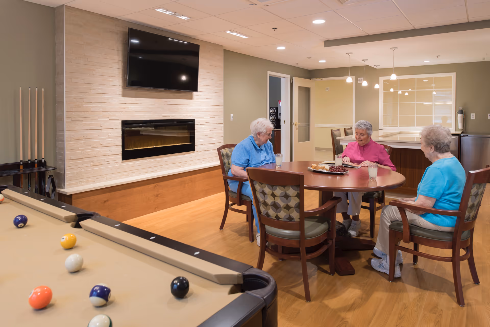 Three elderly women sitting around a round wooden table in a common area, with drinks and a plate of snacks on the table. In the foreground, there is a pool table with balls arranged on it. The room has a mounted TV above a modern electric fireplace on a beige tiled wall, and a rack with pool cues is visible. The space is well-lit with ceiling lights and pendant lamps.