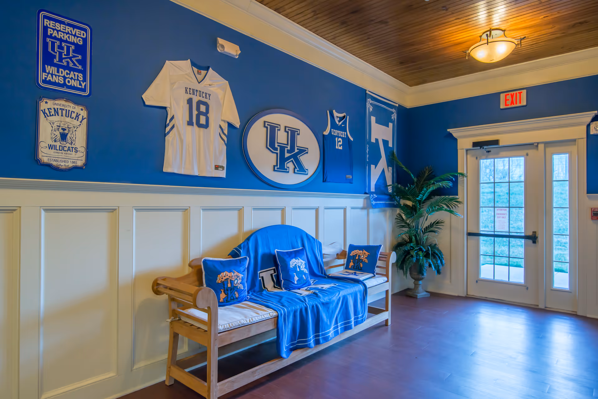 Interior hallway with a wooden bench covered by a blue University of Kentucky blanket and matching pillows. The wall behind the bench is decorated with University of Kentucky sports memorabilia including jerseys, banners, and signs. There is a potted plant near a glass door with an exit sign above it.