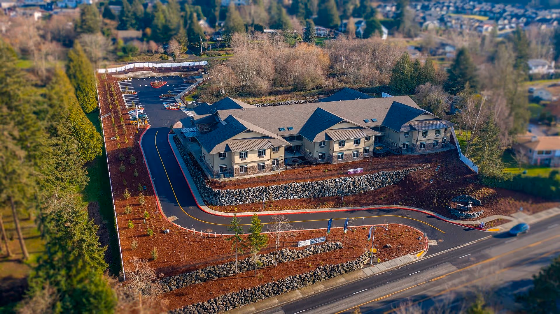 Aerial view of Fieldstone Memory Care Puyallup facility showing a large two-story building surrounded by landscaped grounds with trees, shrubs, and rock retaining walls. The building has multiple roof peaks and skylights. A curved driveway leads to a parking area with several parking spaces. The surrounding area includes residential neighborhoods and wooded areas.
