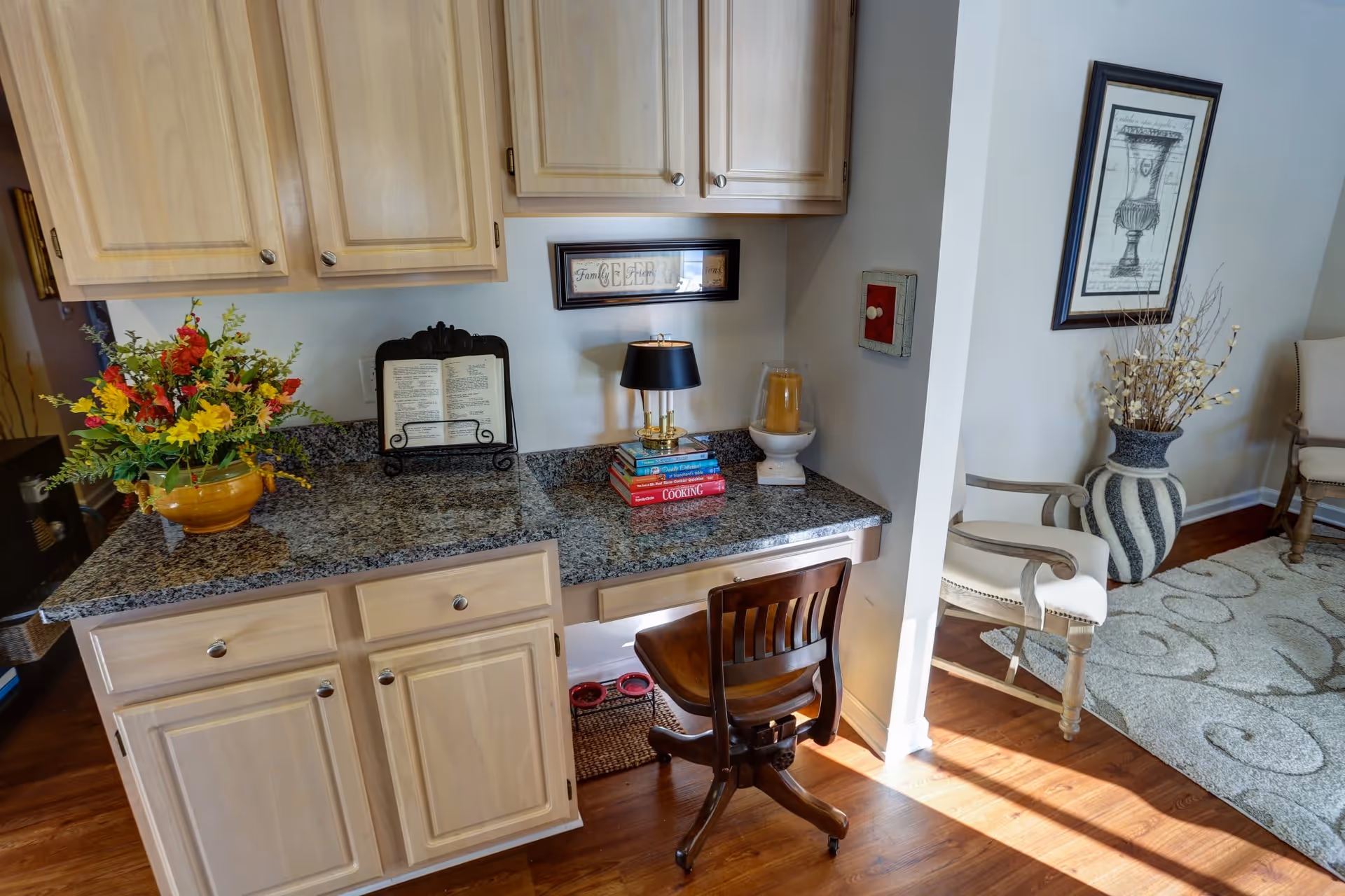 A cozy corner workspace with a granite countertop, light wood cabinets, a wooden chair, and decorative items including a flower vase, an open book on a stand, a small lamp on a stack of books, and a candle holder. Adjacent to the workspace is a living area with a patterned rug, upholstered chairs, a large decorative vase with dried branches, and framed artwork on the wall.