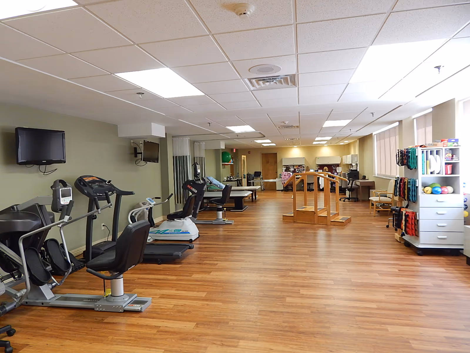 A spacious rehabilitation or physical therapy room with exercise equipment including stationary bikes and treadmills on the left side, wooden handrails in the center for walking practice, and shelves with therapy supplies on the right. The room has wood flooring, beige walls, and ceiling lights.