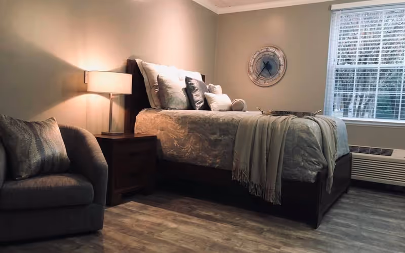 A cozy bedroom featuring a neatly made bed with multiple pillows and a throw blanket. Next to the bed is a wooden nightstand with a modern lamp. A comfortable armchair with a decorative pillow is positioned nearby. A large window with blinds allows natural light to enter the room, and a round wall clock hangs on the wall above the bed. The floor is covered with wood-style flooring.