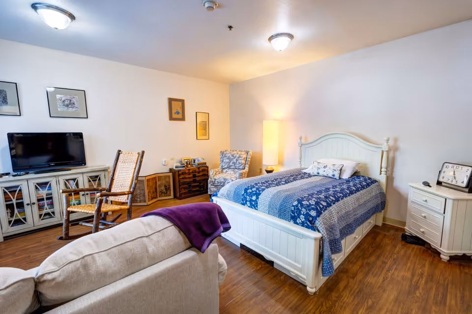 A cozy bedroom with a white wooden bed featuring a blue patterned quilt and pillows. Next to the bed is a white nightstand with a clock and a lamp. There is a cushioned armchair with a floral pattern and a tall floor lamp in the corner. A wooden rocking chair and a TV on a cabinet with glass doors are also visible. The room has wooden flooring and white walls with framed pictures hanging.
