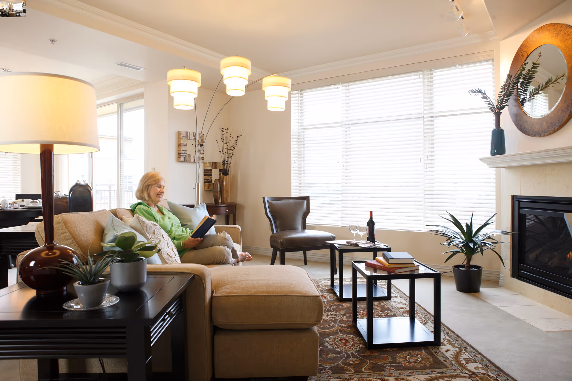 Bright, furnished living room with a person reading on a sofa, coffee tables, plants, and a fireplace.