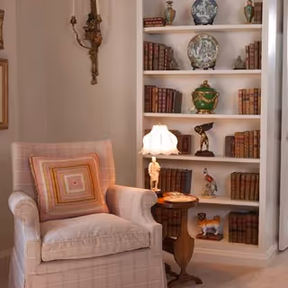 Plush upholstered armchair with a decorative pillow next to a small side table and lamp in front of a built-in bookshelf filled with books and decorative objects.