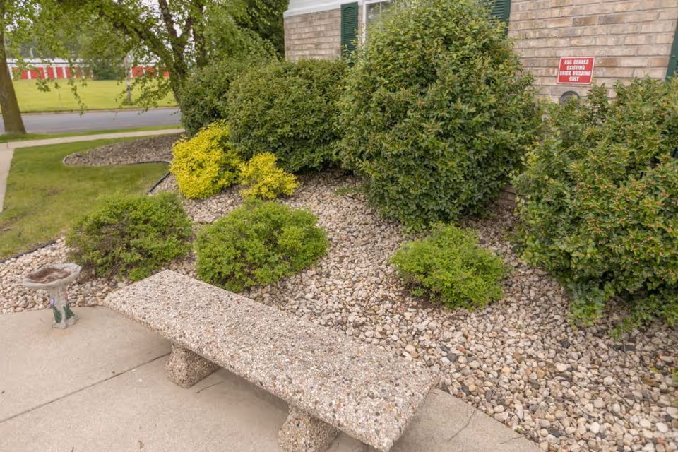 Concrete bench beside a landscaped rock bed with small shrubs in front of a brick building facade.