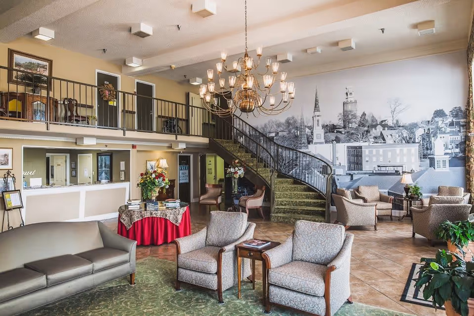 A spacious senior living facility lobby with comfortable seating including sofas and armchairs arranged around small tables. A large chandelier hangs from the ceiling. There is a staircase with a green carpet leading to an upper level with a railing. The back wall features a large black and white mural of a townscape. A reception desk is visible on the left side with flowers and books on a round table covered with a red tablecloth in front of it.