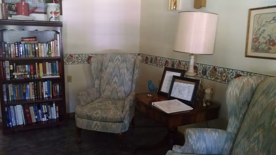 A cozy corner of a room featuring two patterned upholstered armchairs, a wooden side table with a lamp, framed pictures, and a small decorative bird. To the left, there is a tall wooden bookshelf filled with books. The walls have a decorative floral border and a framed artwork.