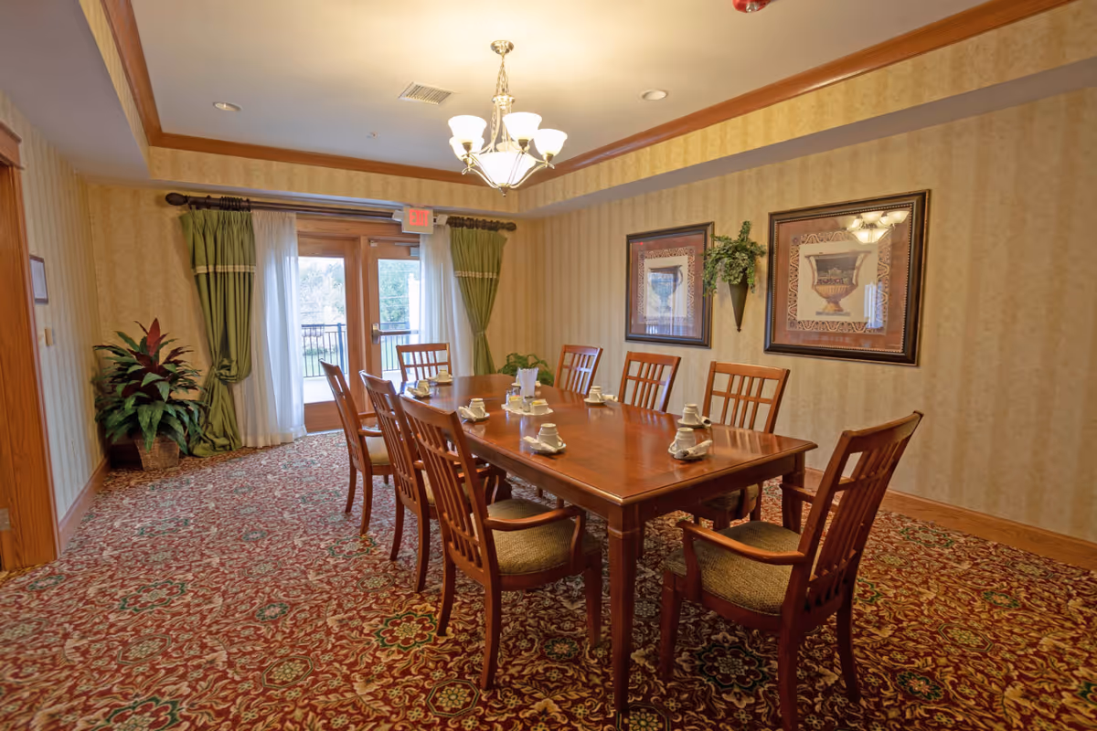 A dining room with a long wooden table set with eight chairs. Each place setting has a cup and saucer with a napkin. The room has patterned carpet, beige wallpaper, two framed pictures on the wall, a chandelier hanging from the ceiling, and a glass door with green curtains leading outside.